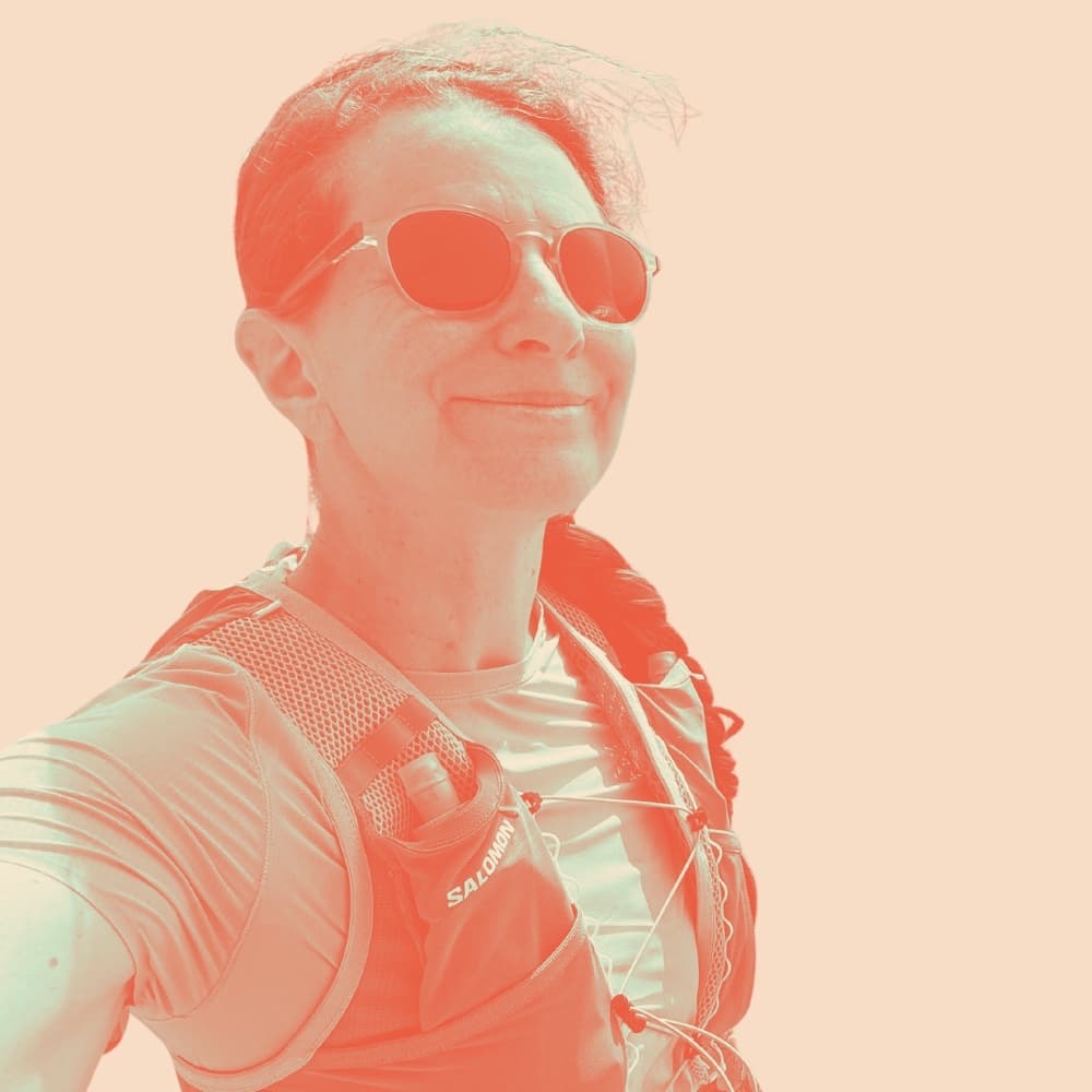 Gwen smiling in sunglasses and a running vest, arm outstretched to take the photo, against a plain peach background.